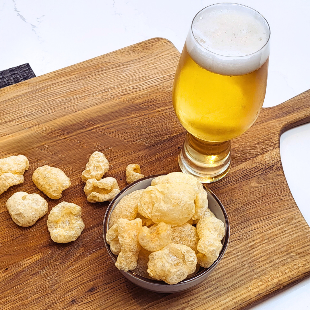 A bowl of Made for Drink's Pork Chicharrones on a wooden serving board with a pint of beer standing next to the bowl and loose pork chicharrones scattered around.