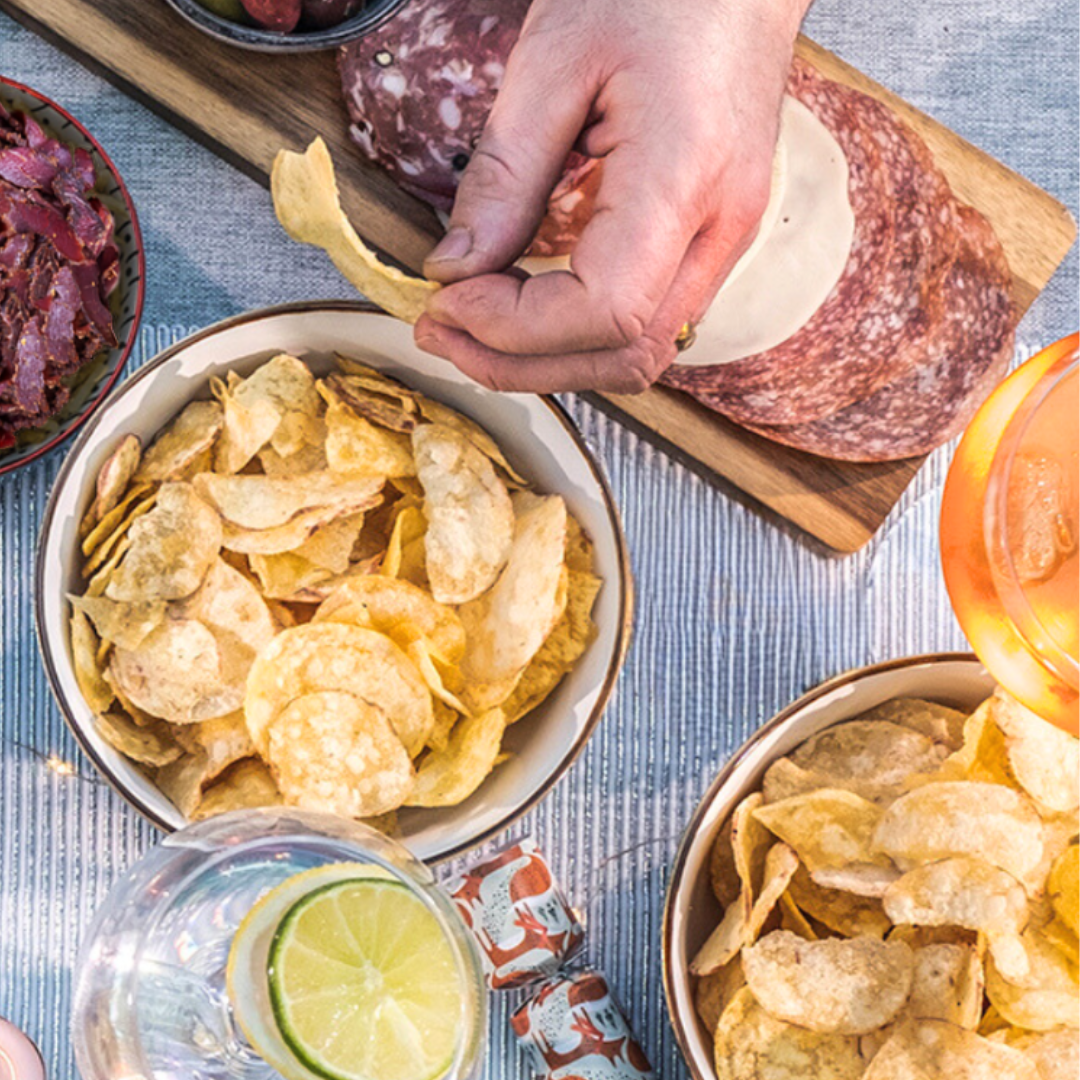 A birds eye view of a dinner table with a grey and silver tablecloth and two bowls of Made For Drink's Dorset Sea Salt Crisps, a charcuterie board of meats and cheese and glasses of gin and tonic and aperol spritz.