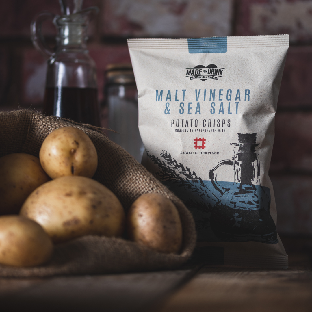 A pack of Made for Drink Malt Vinegar and Sea Sal crisps standing on a wooden table behind a pile of fresh potatoes in a brown hessian sack. In the bacground you can just see a small jug of malt vinegar and a salt shaker.