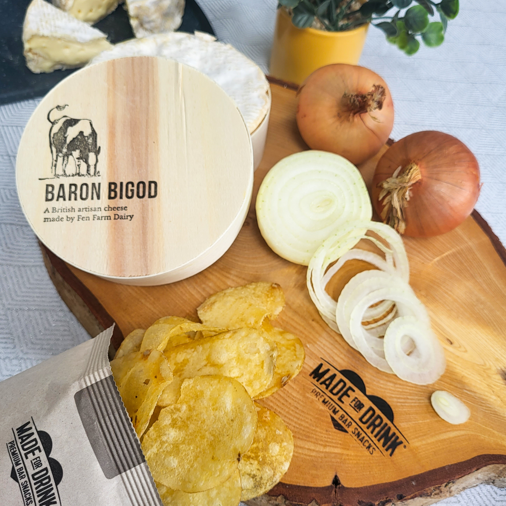 An open packet of Made For Drink's Baron Bigod Cheese & Onion crisps lying across a wooden chopping board, with chopped and whole white onions and a round pack of Baron Bigod cheese from Fen Farm Dairy. You can see slices of brie-style cheese in the background.