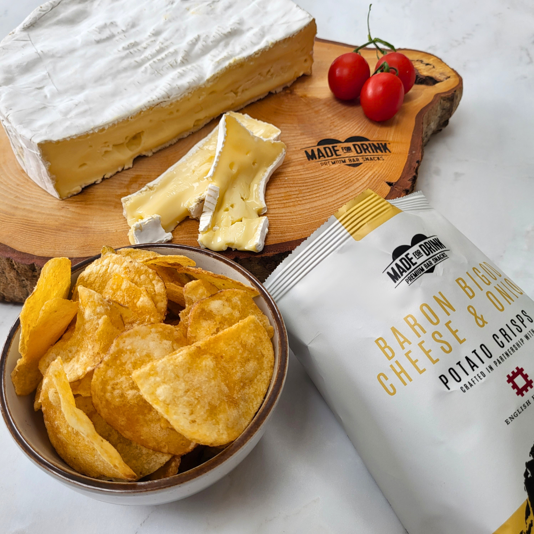 An open packet of Made For Drink's Baron Bigod Cheese & Onion crisps next to a bowl full of the crisps. In the background you can see a wooden chopping board with a large wheel with Baron Bigod brie-style cheese on. It's been sliced across to real the yellow creamy inside and two slices of the cheese are laying on top of each other in the foreground with 3 cherry tomatoes as decoration.