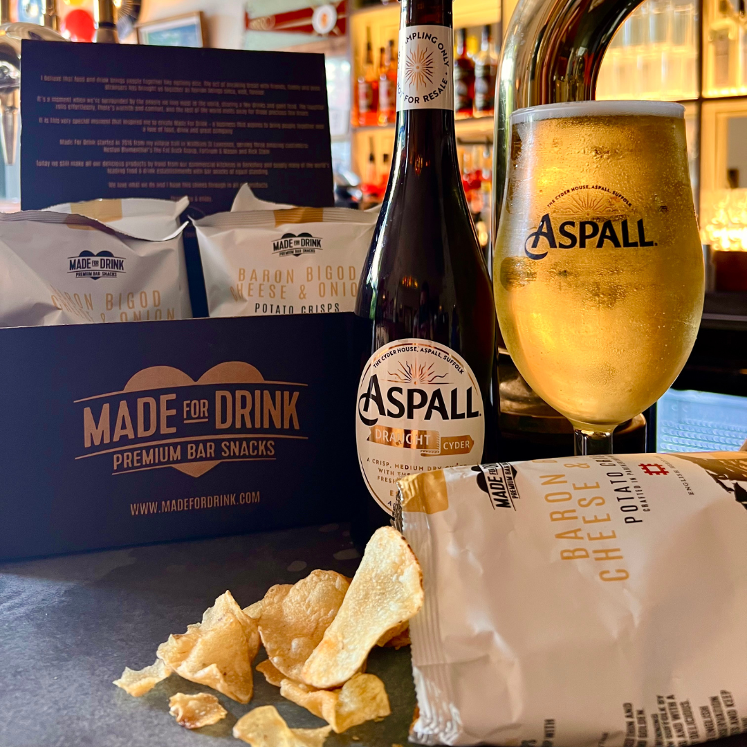 An open packet of Made For Drink's Baron Bigod Cheese & Onion crisps in front of a pint and bottle of cold Aspall Cyder. In the background you can see a branded black An open packet of Made For Drink box of 4 more packets of Baron Bigod Cheese & Onion crisps.