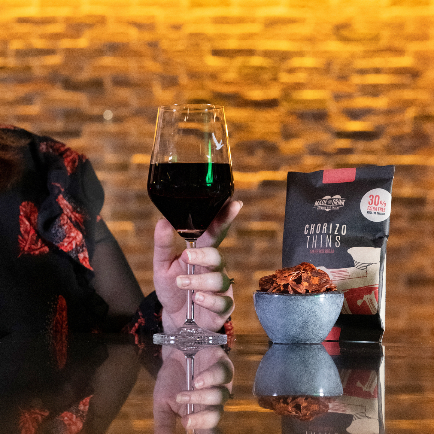 A bowl of Made For Drink's Chorizo Thins on a bar with a pack standing behind and a woman's hand holding a glass of red wine