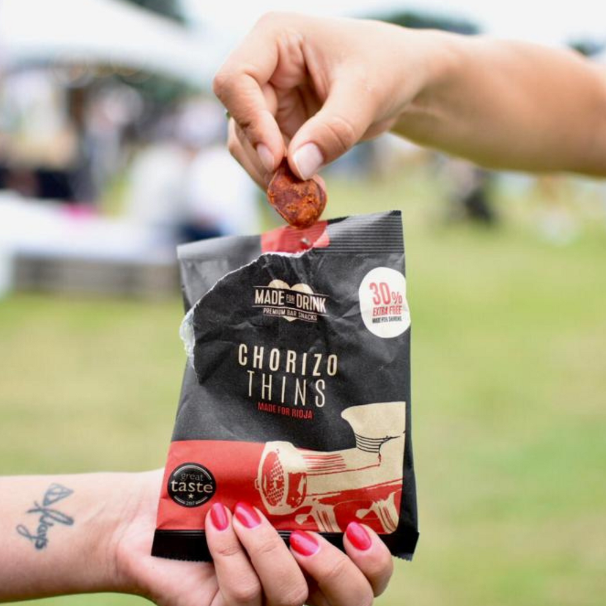 A hand holding an open pack of Made For Drink's Chorizo Thins and another hand reaching into the pack and taking out a handful of chorizo