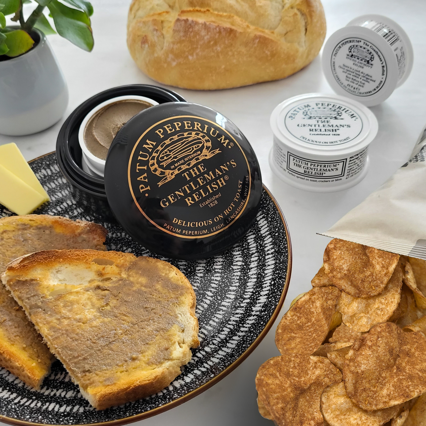 3 pots of Patum Peperium's The Gentleman's Relish, on half opened sowing he paste inside. Rustic bread in the foreground spread with butter and The Gentleman's Relish paste and an open packet of Made For Drink's The Gentleman's Relish potato crisps. to the side.