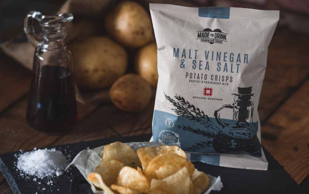 A 40g packet of Made For Drink's Malt Vinegar & Sea Salt crisps standing on a slate plates with a pile of crisps and sea salt in front of it, and a small just of malt vinegar and stack of unpeeled whole potatoes in the background.