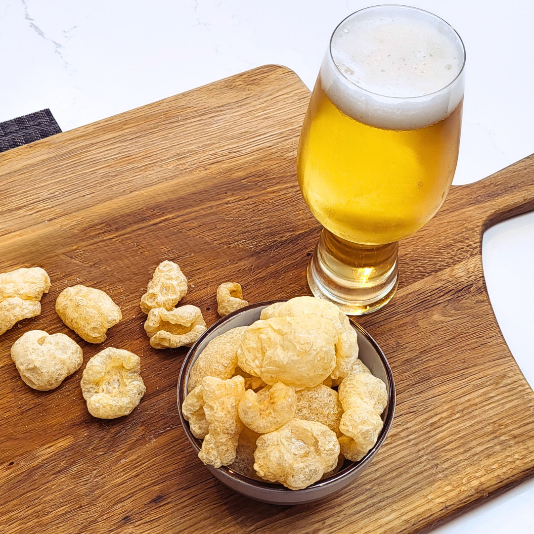 A bowl of Made for Drink's Pork Chicharrones on a wooden serving board with a pint of beer standing next to the bowl and loose pork chicharrones scattered around. 