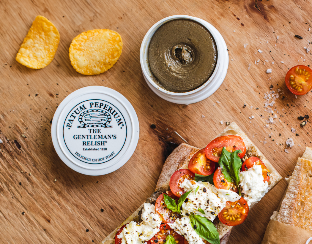 An open pot of The Gentleman's Relish next to a baguette topped The Gentleman's Relish paste, mozzarella, cherry tomatoes and basil and a couple of crisps scattered to the side.